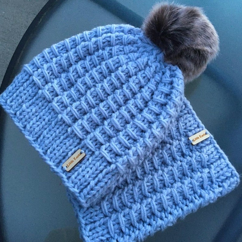 May include: A light blue knitted hat with a brown faux fur pom pom. The hat has a ribbed texture and two wooden tags that say "Little Knitter".