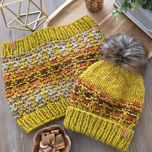 May include: Mustard yellow knitted hat and neck warmer set. The hat has a faux fur pom-pom and a brown, orange, and white stripe design. The neck warmer has a similar stripe pattern. Both items have a leather tag.