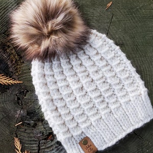 May include: A white knitted beanie with a fluffy brown pom pom on top. The beanie has a textured knit pattern and a brown leather tag with the text "Urban Chic" on it.