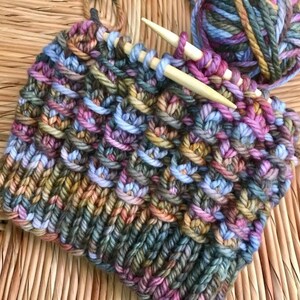 May include: Close-up of a colorful knitted fabric with a multi-colored yarn ball and knitting needles. The fabric is a mix of blues, greens, browns, pinks, and oranges.