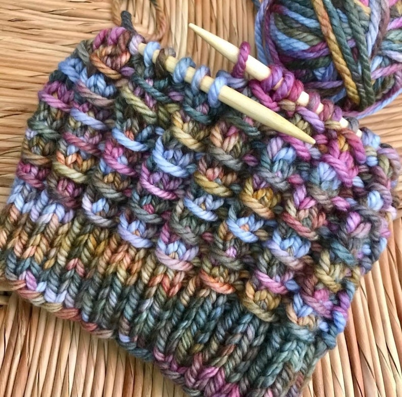 May include: Close-up of a colorful knitted fabric with a multi-colored yarn ball and knitting needles. The fabric is a mix of blues, greens, browns, pinks, and oranges.