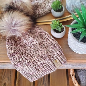 May include: A pink and white knitted beanie with a fluffy brown and white pom pom. The beanie has a leather tag with the text "White Oak Knits" on it.