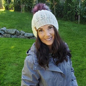 May include: A woman wearing a white knit beanie with a pink pom pom and a gray jacket. She is smiling and looking at the camera.