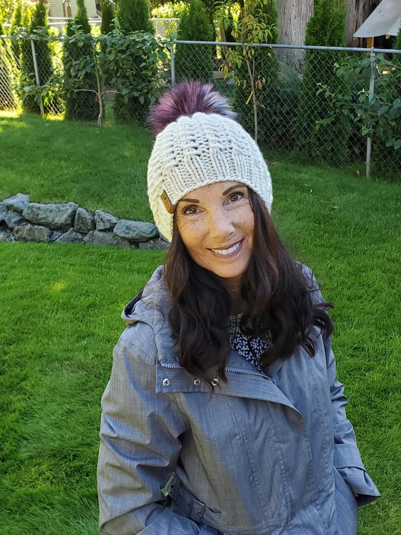 May include: A woman wearing a white knit beanie with a pink pom pom and a gray jacket. She is smiling and looking at the camera.