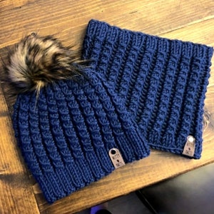 May include: A navy blue knitted hat with a faux fur pom pom and a matching navy blue knitted neck warmer. Both items have a brown leather tag with the text "Knotty Knits".