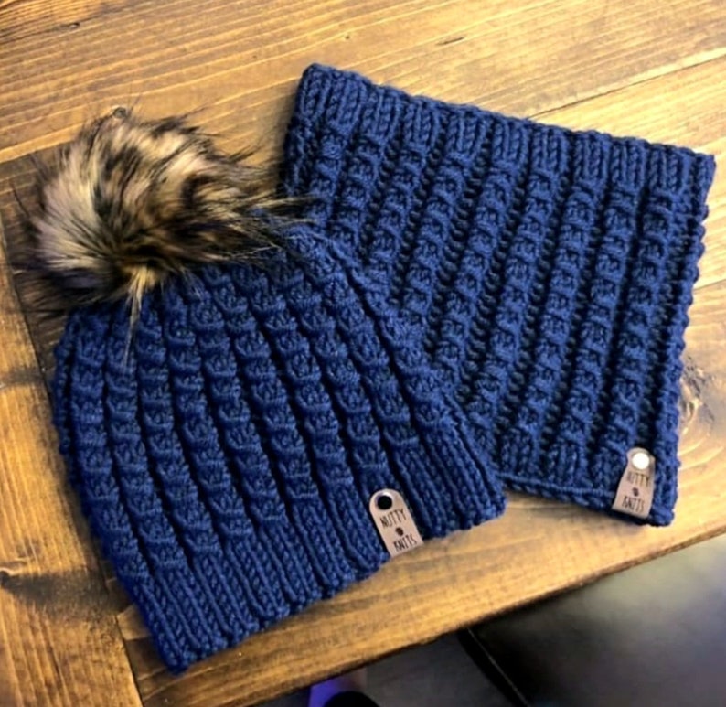 May include: A navy blue knitted hat with a faux fur pom pom and a matching navy blue knitted neck warmer. Both items have a brown leather tag with the text "Knotty Knits".