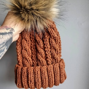 May include: A brown knitted beanie with a large brown and white faux fur pom pom on top.