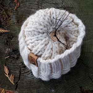 May include: A white knitted beanie with a brown leather tag that says "Cozy Creations" and a brown wooden button with the text "Handmade with love".