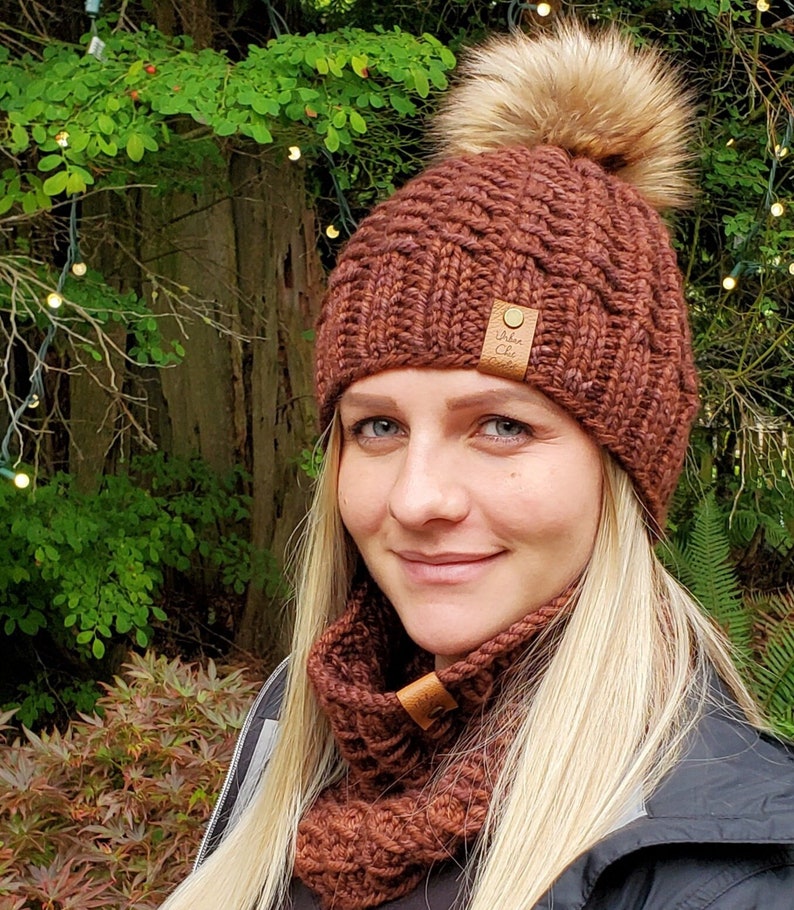 May include: A brown knitted hat with a faux fur pom pom and a matching brown knitted neck warmer. The hat has a brown leather tag with the text "Urban Chic" on it.