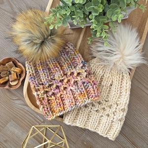 May include: Two knitted beanies with faux fur pom-poms. One beanie is cream-colored, and the other is multi-colored with shades of pink, yellow, and blue. The beanies are displayed on a wooden tray with a small plant and decorative items.