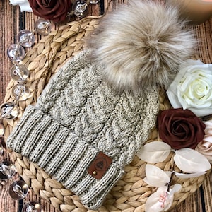 May include: A beige knitted beanie with a cable knit pattern and a faux fur pom pom. The beanie has a brown leather patch with the text "Celtic Knot Company" on it.