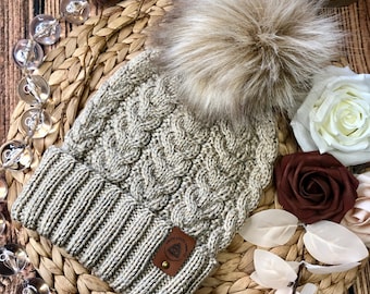 May include: A beige knitted beanie with a cable knit pattern and a faux fur pom pom. The beanie has a brown leather patch with the text "Celtic Knot Company" on it.
