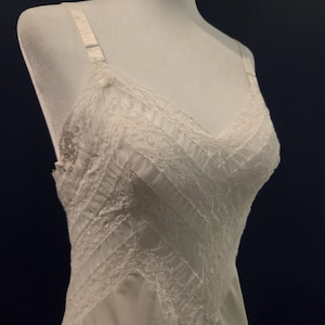 May include: A white vintage slip dress with delicate lace detailing. The dress features a V-neckline and adjustable straps. The bodice has a textured, layered design, and the skirt flows smoothly. The slip is displayed on a mannequin against a dark background.