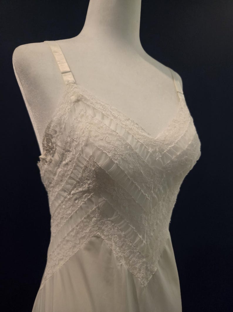 May include: A white vintage slip dress with delicate lace detailing. The dress features a V-neckline and adjustable straps. The bodice has a textured, layered design, and the skirt flows smoothly. The slip is displayed on a mannequin against a dark background.