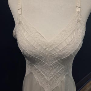 May include: A white slip dress with a V-neckline and delicate lace trim. The dress features adjustable straps and a flowing, lightweight design. The bodice has a chevron pattern with pleated details.