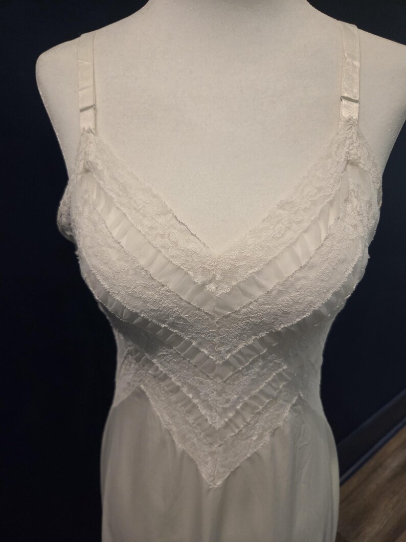 May include: A white slip dress with a V-neckline and delicate lace trim. The dress features adjustable straps and a flowing, lightweight design. The bodice has a chevron pattern with pleated details.