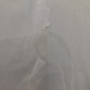 May include: A close-up of a light gray fabric with a small tear. The fabric appears to be smooth and possibly a lightweight material. The tear is located near the center of the image.