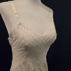 May include: A white slip dress on a mannequin. The dress features a V-neckline and delicate lace trim along the neckline and hem. The bodice has a textured design with vertical and diagonal panels. Adjustable straps complete the design.