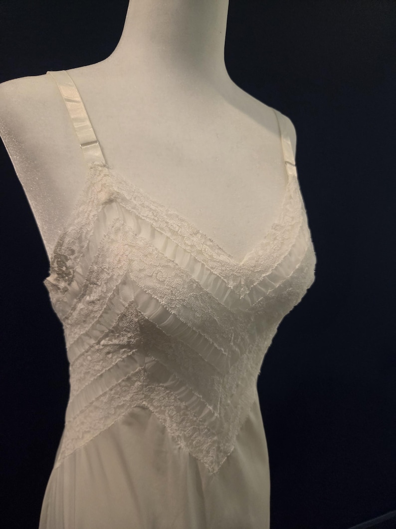 May include: A white slip dress on a mannequin. The dress features a V-neckline and delicate lace trim along the neckline and hem. The bodice has a textured design with vertical and diagonal panels. Adjustable straps complete the design.
