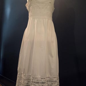 May include: A white slip dress with delicate lace trim. The dress features spaghetti straps and a fitted bodice with lace detailing. The hemline is adorned with a wide band of intricate lace, creating a vintage aesthetic.