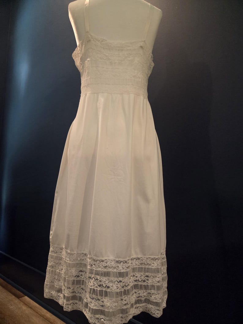 May include: A white slip dress with delicate lace trim. The dress features spaghetti straps and a fitted bodice with lace detailing. The hemline is adorned with a wide band of intricate lace, creating a vintage aesthetic.