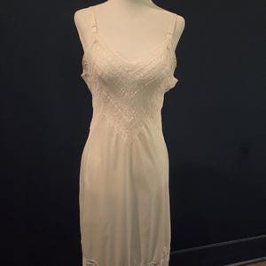 May include: A white slip dress with adjustable straps and a V-neckline. The bodice features a detailed lace design, and the hemline is trimmed with delicate lace. The dress is displayed on a white mannequin against a dark blue background.