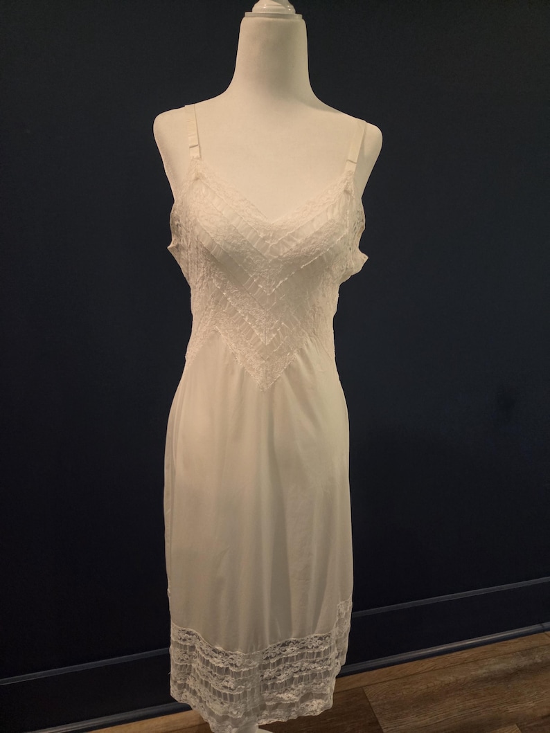 May include: A white slip dress with adjustable straps and a V-neckline. The bodice features a detailed lace design, and the hemline is trimmed with delicate lace. The dress is displayed on a white mannequin against a dark blue background.