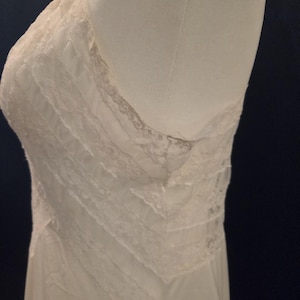 May include: Ivory-colored slip dress with delicate lace trim. The dress features a V-neckline and layered bodice with intricate lace detailing. The fabric appears soft and flowing, suggesting a vintage or romantic style.