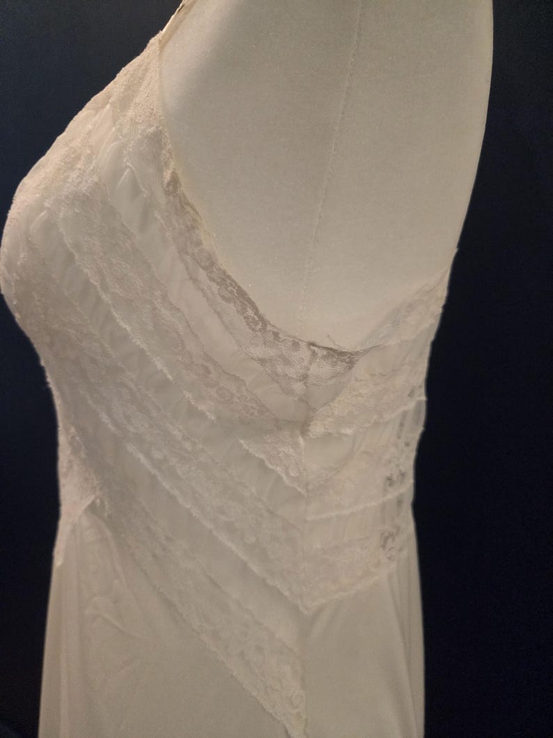May include: Ivory-colored slip dress with delicate lace trim. The dress features a V-neckline and layered bodice with intricate lace detailing. The fabric appears soft and flowing, suggesting a vintage or romantic style.