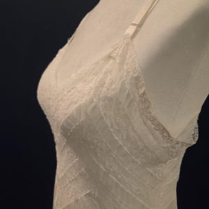 May include: Ivory-colored lingerie with delicate lace detailing. The slip features a V-neckline and adjustable straps. The design includes horizontal lace accents across the bodice, creating a layered effect. The fabric appears soft and lightweight, suitable for sleepwear or undergarments.