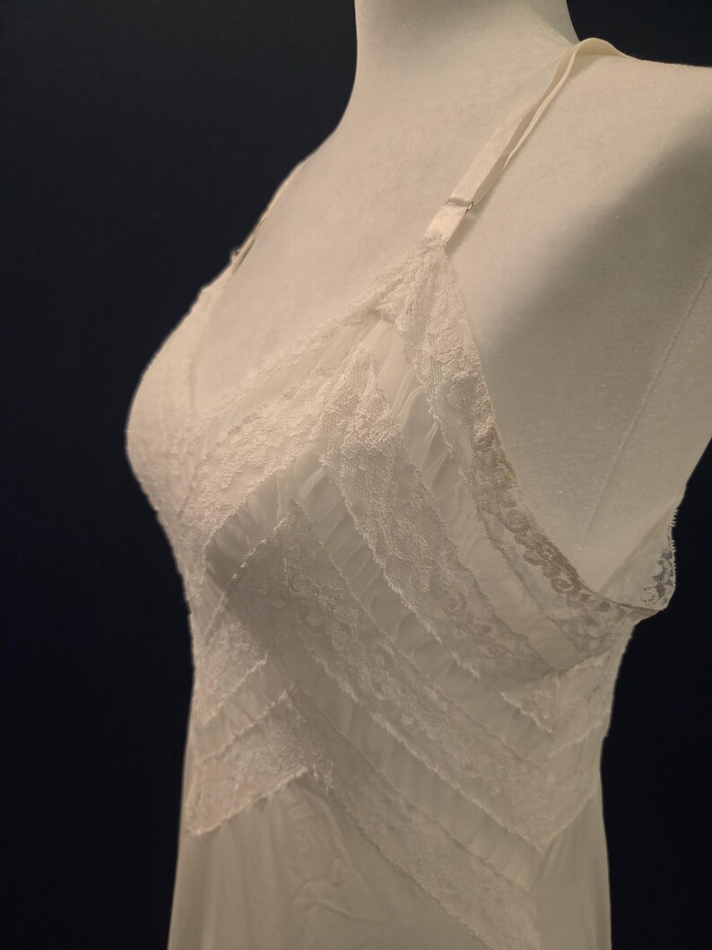 May include: Ivory-colored lingerie with delicate lace detailing. The slip features a V-neckline and adjustable straps. The design includes horizontal lace accents across the bodice, creating a layered effect. The fabric appears soft and lightweight, suitable for sleepwear or undergarments.