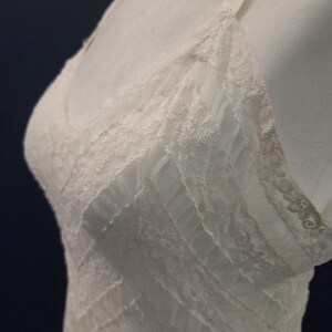 May include: A white lingerie slip with delicate lace trim and thin straps. The slip features a V-neckline and a layered design, creating a textured appearance. The garment is displayed on a mannequin.