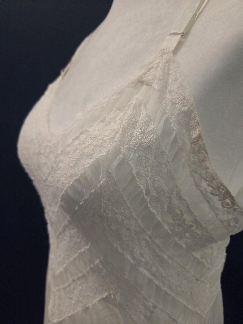 May include: A white lingerie slip with delicate lace trim and thin straps. The slip features a V-neckline and a layered design, creating a textured appearance. The garment is displayed on a mannequin.
