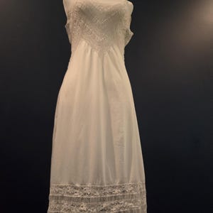 May include: A white slip dress displayed on a mannequin. The dress features a detailed lace design on the bodice and hem. The dress is sleeveless and has a flowing, elegant silhouette, perfect for vintage or bridal wear.