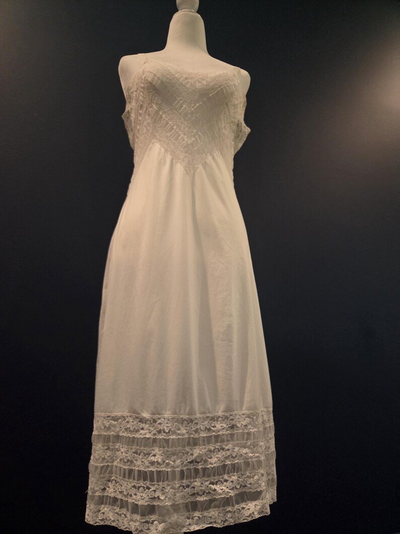 May include: A white slip dress displayed on a mannequin. The dress features a detailed lace design on the bodice and hem. The dress is sleeveless and has a flowing, elegant silhouette, perfect for vintage or bridal wear.