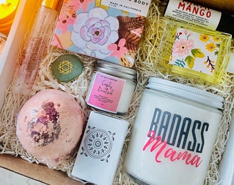 May include: A gift box filled with various self-care items, including a bath and body soap, a candle, a bath bomb, a bottle of essential oil, and a bottle of body spray. The box is decorated with shredded paper and a small wooden sign that reads "Badass Mama".