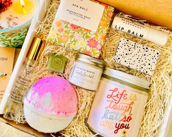 May include: A gift box filled with self-care items, including a bath bomb, candle, soap, lip balm, and a small jar of lotion. The candle has the text "Life is tough but so are you" on the label. The box is filled with shredded paper.