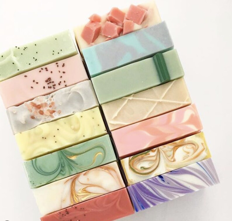 May include: A stack of 15 handmade soap bars in various colors and patterns, including green, pink, purple, and white. The soaps have different textures and designs, such as swirls, speckles, and stripes.