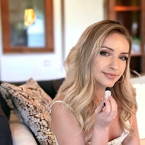 May include: A woman with long blonde hair is sitting on a couch and holding a small white tube in her hand. She is wearing a white tank top and has a neutral expression on her face.