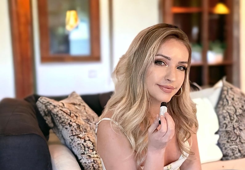 May include: A woman with long blonde hair is sitting on a couch and holding a small white tube in her hand. She is wearing a white tank top and has a neutral expression on her face.