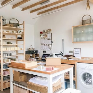 May include: A white work table with a wooden top, filled with boxes of handmade soap bars. The table is in a room with a white washing machine, a wooden shelf with soap bars, and a wooden table with a laptop and a printer.