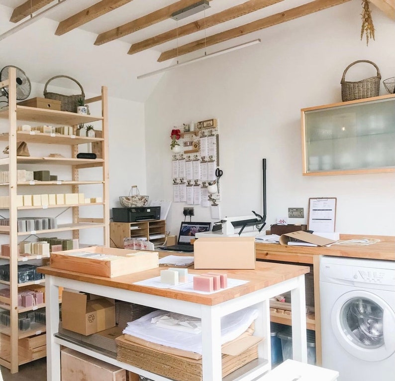 May include: A white work table with a wooden top, filled with boxes of handmade soap bars. The table is in a room with a white washing machine, a wooden shelf with soap bars, and a wooden table with a laptop and a printer.