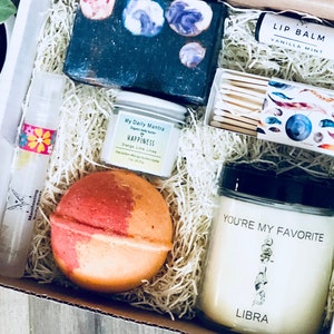 May include: A gift box filled with self-care items, including a bath bomb, a candle, a bar of soap, a lip balm, a small jar of body butter, and a box of matches. The candle has the text "You're My Favorite Libra" on the front. The bath bomb is orange and red. The soap is black with a galaxy print. The lip balm is black with white text that says "Lip Balm Vanilla Mint". The body butter is white with the text "My Daily Mantra Happiness Orange Lime Litsea". The box of matches has a galaxy print with feathers.