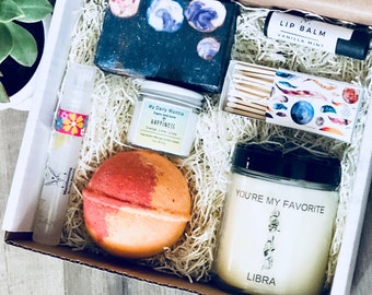 Può includere: Una scatola regalo piena di articoli per la cura di sé, tra cui una bomba da bagno, una candela, un sapone, un balsamo per le labbra, un piccolo barattolo di burro per il corpo e una scatola di fiammiferi. La candela ha il testo "You're My Favorite Libra" sul davanti. La bomba da bagno è arancione e rossa. Il sapone è nero con una stampa galattica. Il balsamo per le labbra è nero con testo bianco che dice "Lip Balm Vanilla Mint". Il burro per il corpo è bianco con il testo "My Daily Mantra Happiness Orange Lime Litsea". La scatola di fiammiferi ha una stampa galattica con piume.