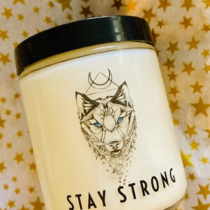 May include: White candle with a black lid and a wolf graphic. The wolf has blue eyes and is surrounded by a geometric design. The text "STAY STRONG" is printed on the front of the candle.
