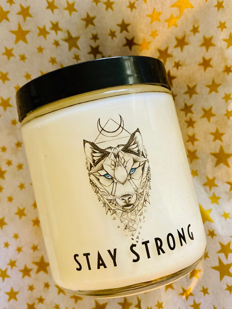 May include: White candle with a black lid and a wolf graphic. The wolf has blue eyes and is surrounded by a geometric design. The text "STAY STRONG" is printed on the front of the candle.