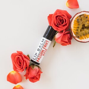 K&ouml;nnte beinhalten: Eine wei&szlig;e Tube Lippenbalsam mit schwarzem Text, der "Lip Balm Passion Fruit Rose" lautet. Die Tube ist von roten Rosenbl&auml;ttern und einer Passionsfrucht umgeben.