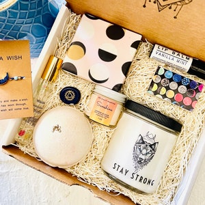 May include: A gift box filled with self-care items, including a bath bomb, a candle, a lip balm, a small bottle of essential oil, and a small jar of body scrub. The box is decorated with shredded paper and a small card that says "Make a Wish".