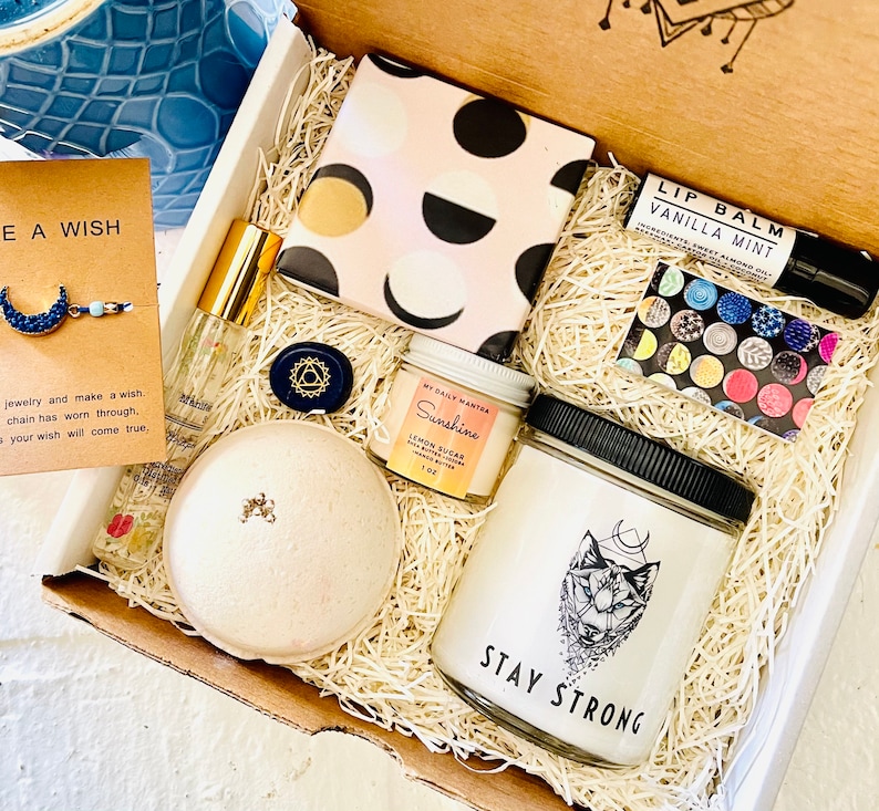 May include: A gift box filled with self-care items, including a bath bomb, a candle, a lip balm, a small bottle of essential oil, and a small jar of body scrub. The box is decorated with shredded paper and a small card that says "Make a Wish".