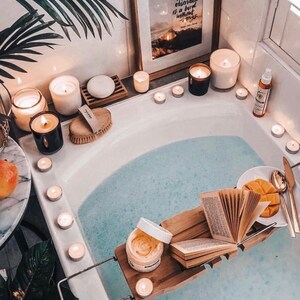 May include: A white bathtub filled with blue water, surrounded by lit candles and a wooden bath tray with a book, a bowl of fruit, and a jar of bath products. The text on the framed print reads "A traveler without observation is a bird without wings."